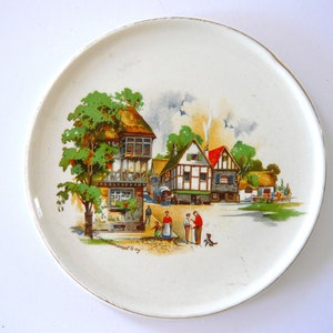 May include: A white ceramic plate with a painted scene of a village with houses, trees, and people. The plate has a gold rim.
