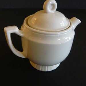 May include: Cream-colored ceramic teapot with a lid and a loop handle. The teapot has a spout and a handle on the side. The base of the teapot has a decorative, ridged design. The teapot is sitting on a dark surface.