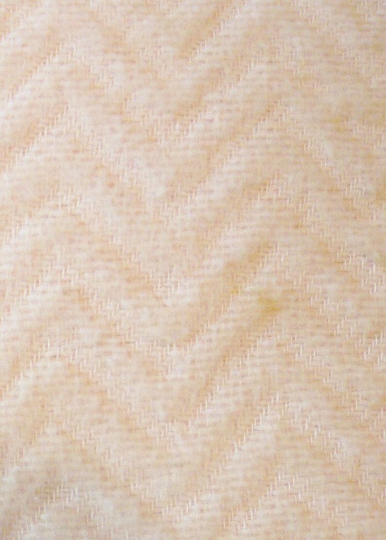 May include: Close-up of a textured fabric with a diagonal herringbone pattern. The fabric has a light beige color with subtle variations in tone, creating a soft, inviting appearance. The weave is tight and detailed.