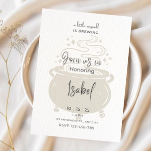 May include: A white invitation card with a whimsical design, featuring a cauldron and the text "a little wizard is brewing." The card invites guests to honor Isabel, with date, time, and RSVP information.
