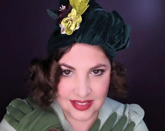 May include: A woman wearing a green velvet turban with a yellow and brown flower embellishment. She is wearing green gloves and a light green blouse.