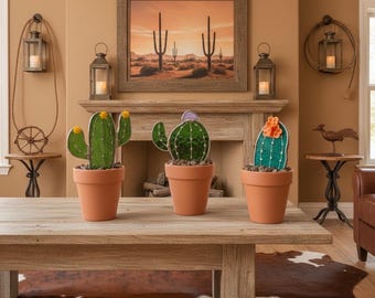 May include: Three decorative cactus plants in terracotta pots sit on a wooden table. The cacti are green with yellow and orange accents. A framed desert landscape painting hangs above a fireplace. The room has a rustic, southwestern theme.