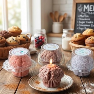 Pode incluir: Três velas em formato de muffin em recipientes de plástico transparente, uma rosa, uma castanha e uma roxa clara. Uma quarta vela castanha em formato de muffin está sobre um prato pequeno com um pavio aceso.