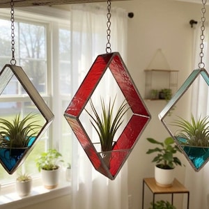 May include: Three hanging geometric planters with air plants. The planters are diamond-shaped with stained glass in red, teal, and clear. Each planter has a teal or blue base. The plants have long green leaves.