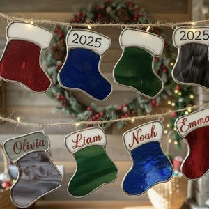 May include: A collection of stained glass Christmas stocking ornaments in various colors, including red, blue, green, and black, strung on a twine garland. Some stockings have names like "Olivia," "Liam," "Noah," and "Emma," while others display the year "2025."