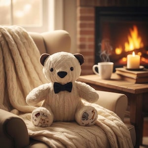 Pode incluir: Um urso de pelúcia creme com uma gravata borboleta preta está sentado em um cobertor de malha creme. O urso tem orelhas marrons e detalhes bordados. A cena inclui uma lareira, uma vela acesa e uma xícara de café, criando uma atmosfera acolhedora.