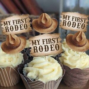May include: Four cupcakes decorated with brown cowboy hats and signs that say "My First Rodeo". The cupcakes are topped with white frosting and have brown cupcake liners with a wood grain pattern.