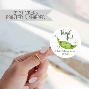 May include: A round 2-inch sticker featuring a green pea pod illustration with two smiling peas inside. The text reads Thank You!, Sabrina's baby shower, and 03/24/24. The design includes small floral accents and a thin heart-shaped vine.