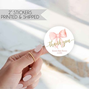 May include: Round thank you sticker with a pink watercolor bow and gold lettering. The sticker says "thank you" with "Kristin's Baby Shower 2.2.2022" below.