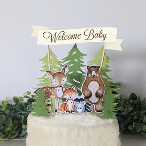 K&ouml;nnte beinhalten: Ein wei&szlig;er Kuchen mit einem Waldtier-Motiv. Der Kuchen ist mit einem Banner verziert, auf dem "Willkommen Baby" steht, sowie einem B&auml;ren, einem Reh, einem Fuchs und einem Waschb&auml;ren. Der Kuchen ist von gr&uuml;nen Bl&auml;ttern umgeben.