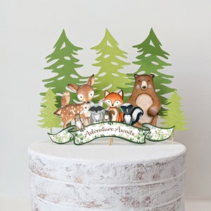 May include: A white cake with a woodland animal themed topper. The topper features a banner that says "Adventure Awaits" with a deer, fox, raccoon, bear, owl, and skunk.