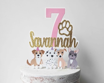 Puede incluir: Un pastel blanco con un número 7 rosa y un adorno de purpurina dorada 'Savannah'. El pastel está decorado con cinco perros de dibujos animados, cada uno con una raza y un color diferentes. El pastel está sobre una base de madera.