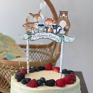 May include: A white cake with buttercream frosting and a woodland animal themed baby shower topper that says "Welcome Baby". The cake is decorated with fresh berries.