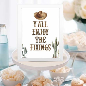 May include: A white framed sign with the text "Y'all Enjoy The Fixings" in brown lettering. A cowboy hat and cactus illustrations are on the sign. The sign is on a white tiered cake stand with blue and white desserts on the table.