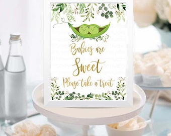 Może przedstawiać: Biały znak z zielonymi liśćmi i dwoma groszkami w strąku, na którym widnieje napis "Babies are Sweet Please take a treat". Znak stoi na białym stojaku na ciasto z różnymi deserami na białym stole.