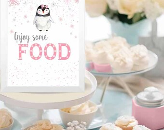 May include: A white sign with a pink penguin illustration and the text "Enjoy some FOOD" in pink letters with white polka dots. The sign is on a white cake stand with a white background and a light blue table with various desserts.