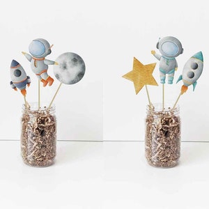May include: Two sets of astronaut themed cake toppers. Each set includes an astronaut, a rocket, a star, and a moon. The toppers are made of cardstock and are perfect for a space-themed party.