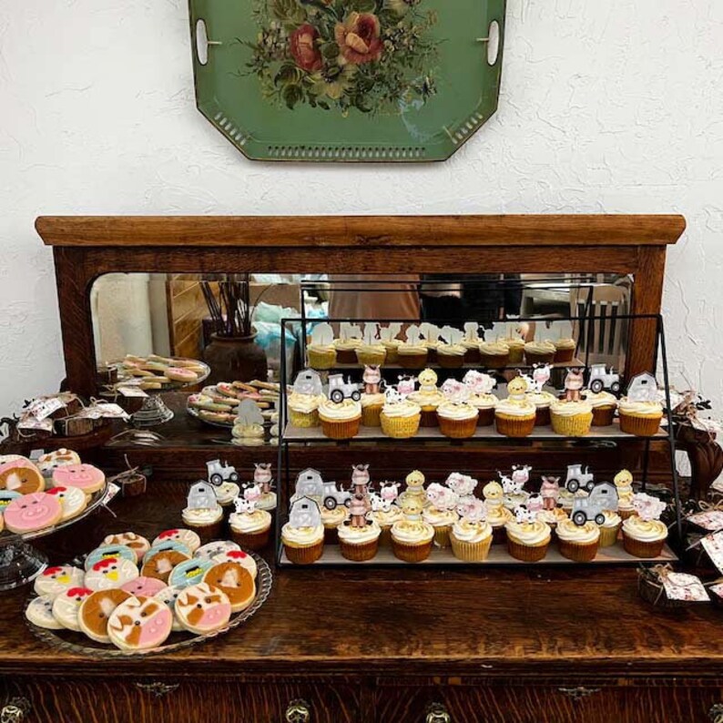 May include: A two-tiered cupcake stand filled with cupcakes decorated with farm animal and tractor toppers. The cupcakes are displayed on a wooden table with a mirror behind them. There are also cookies on the table, decorated with farm animal designs.