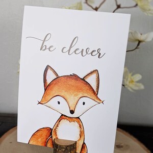 Puede incluir: Una tarjeta blanca con el texto "be clever" en escritura cursiva marr&oacute;n. Un zorro de dibujos animados con pelaje naranja y pecho blanco est&aacute; ilustrado en la tarjeta. La tarjeta est&aacute; apoyada en un peque&ntilde;o tronco de madera.