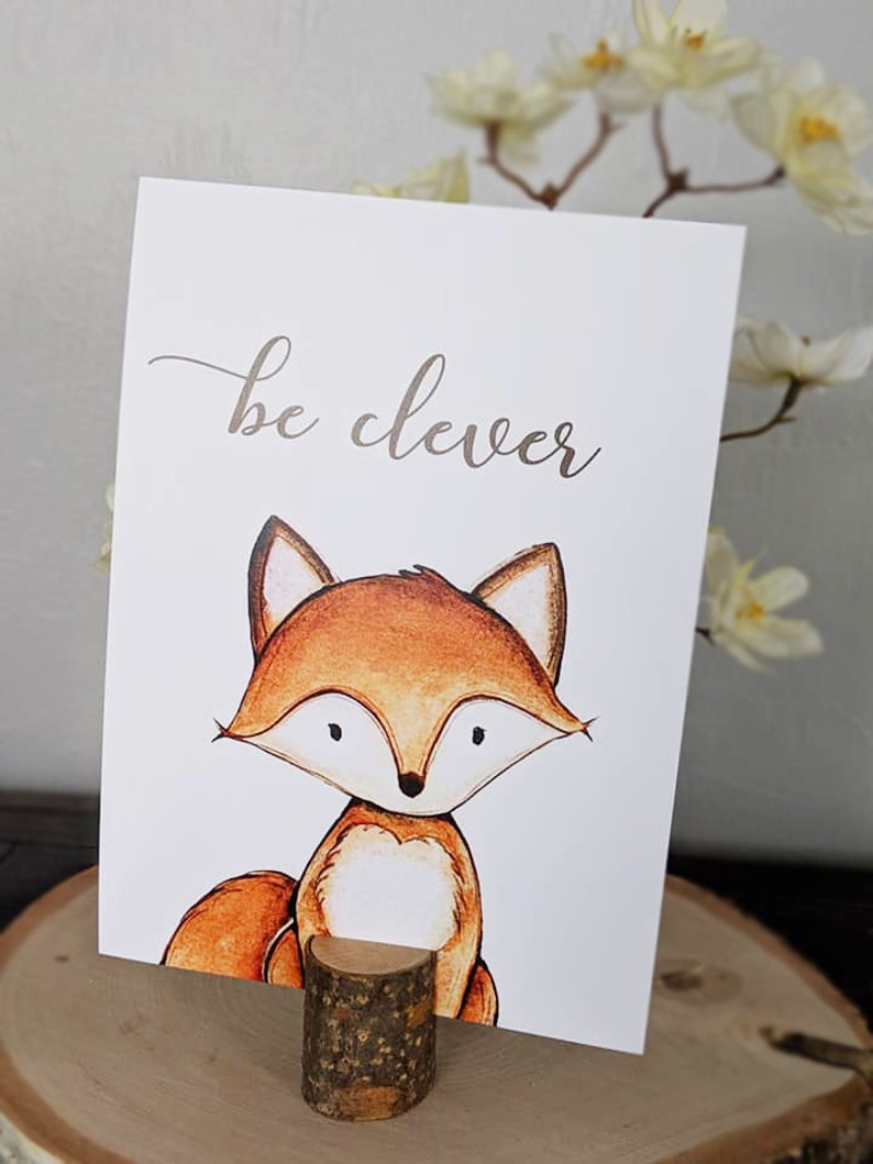Puede incluir: Una tarjeta blanca con el texto "be clever" en escritura cursiva marr&oacute;n. Un zorro de dibujos animados con pelaje naranja y pecho blanco est&aacute; ilustrado en la tarjeta. La tarjeta est&aacute; apoyada en un peque&ntilde;o tronco de madera.