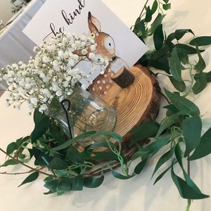 Puede incluir: Una rodaja de madera con un peque&ntilde;o cartel que dice "be kind" y una ilustraci&oacute;n de un ciervo. La rodaja est&aacute; decorada con flores blancas de aliento de beb&eacute; en un frasco de vidrio y una guirnalda de hojas verdes.