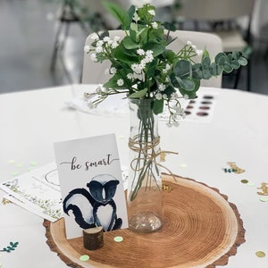 Puede incluir: Una mesa blanca con una rodaja de madera como mantel individual. Un jarr&oacute;n de vidrio transparente con un ramo de flores blancas y hojas verdes est&aacute; en el centro. Una tarjeta blanca con el texto "be smart" y una mofeta de dibujos animados est&aacute; sobre la mesa.