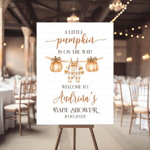 Könnte beinhalten: Ein weißes Schild auf einer Holzstaffelei mit Text und Illustrationen für eine Babyparty. Auf dem Schild steht "A little pumpkin is on the way!" mit Kürbis- und Strampler-Illustrationen. Die Babyparty ist für Audrina und das Datum ist der 10.10.2025.