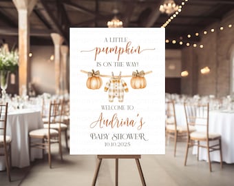 May include: A white rectangular sign on a wooden easel featuring the text A LITTLE pumpkin IS ON THE WAY! with watercolor illustrations of two pumpkins and a baby onesie on a clothesline, plus WELCOME TO Audrina's BABY SHOWER 10.10.2025.