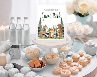 May include: A white sign with a white frame and the text "Please sign the Guest Book" in black. The sign is decorated with a woodland scene featuring a deer, fox, raccoon, bear, and other animals. The sign is on a white cake stand with a white background.