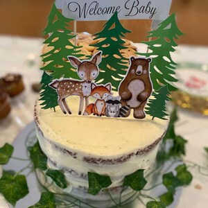 K&ouml;nnte beinhalten: Ein wei&szlig;er, mit Zuckerguss &uuml;berzogener Kuchen, der mit gr&uuml;nen B&auml;umen, einem Reh, einem Fuchs, einem Waschb&auml;ren und einem B&auml;ren verziert ist. Der Kuchen hat ein Banner mit der Aufschrift "Willkommen Baby".