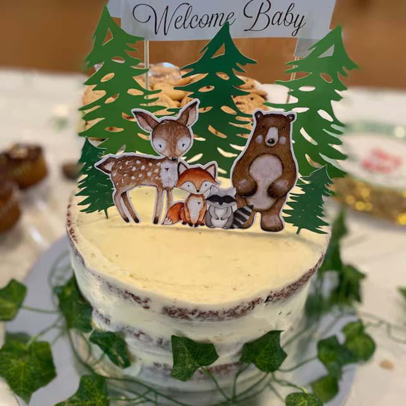 K&ouml;nnte beinhalten: Ein wei&szlig;er, mit Zuckerguss &uuml;berzogener Kuchen, der mit gr&uuml;nen B&auml;umen, einem Reh, einem Fuchs, einem Waschb&auml;ren und einem B&auml;ren verziert ist. Der Kuchen hat ein Banner mit der Aufschrift "Willkommen Baby".