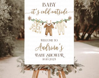 Może przedstawiać: Biały prostokątny znak na drewnianej sztaludze z napisem BABY it's cold outside, WELCOME TO Audrina's BABY SHOWER 10.10.2025. Projekt zawiera sznur na pranie z trzema ubrankami dla niemowląt, gałązkami sosny i płatkami śniegu.