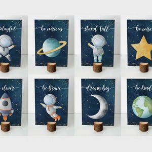 May include: Set of eight watercolor art prints with space-themed illustrations. Each print features a different image, including an astronaut, planet, star, rocket, and moon, with inspirational phrases like "be playful" and "dream big."