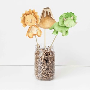 May include: Three watercolor dinosaur party decorations, including a green triceratops, a brown triceratops, and a brown volcano, are displayed in a clear glass jar filled with brown shredded paper.
