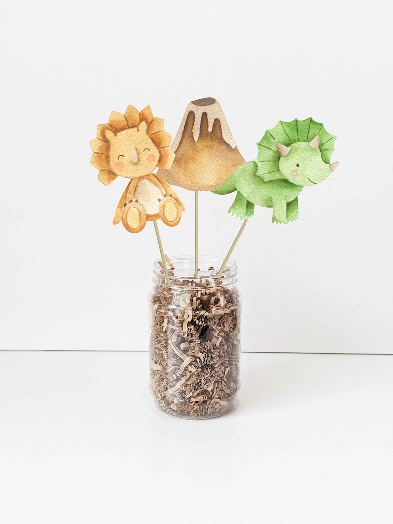 May include: Three watercolor dinosaur party decorations, including a green triceratops, a brown triceratops, and a brown volcano, are displayed in a clear glass jar filled with brown shredded paper.