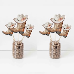 May include: Two glass jars filled with brown shredded paper, each holding three cowboy boot and hat decorations. The boots and hats are light brown with floral accents, and are attached to wooden sticks.