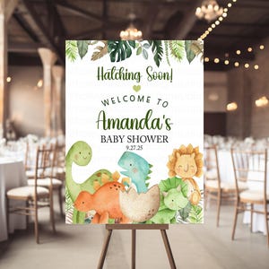 Può includere: Un cartello di benvenuto per un baby shower con illustrazioni di dinosauri e foglie. Il cartello recita "Hatching Soon! Welcome to Amanda's Baby Shower 9.27.25". Il cartello è su un cavalletto di legno in uno spazio eventi decorato.