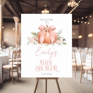 May include: A baby shower welcome sign featuring a watercolor pumpkin with a bow and floral accents. The sign reads "Welcome To Emily's Baby Shower 11.12.2024" in elegant script. The sign is displayed on a wooden easel.