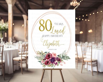 May include: A white sign on a wooden easel with gold text and floral accents. The sign reads "80 Years Loved Happy Birthday Elizabeth August 8, 2020." The text is inside a gold oval frame, with a floral arrangement at the bottom.