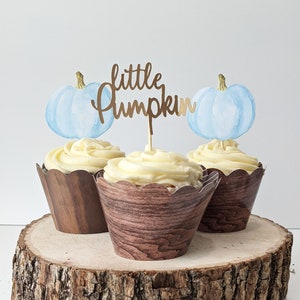 May include: Three cupcakes with light yellow frosting and wooden-look paper wrappers. Each cupcake has a light blue watercolor pumpkin and a gold-colored "little pumpkin" topper. The cupcakes are displayed on a rustic wooden slice.