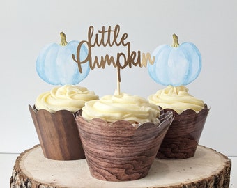 May include: Three cupcakes with light yellow frosting and wooden-look paper wrappers. Each cupcake has a light blue watercolor pumpkin and a gold-colored "little pumpkin" topper. The cupcakes are displayed on a rustic wooden slice.