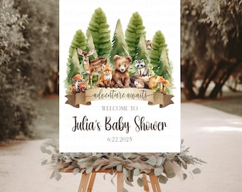May include: A baby shower sign featuring woodland animals and pine trees. The sign reads "adventure awaits" and "Welcome to Julia's Baby Shower" with the date 6.22.2025. The sign is displayed on a wooden easel.