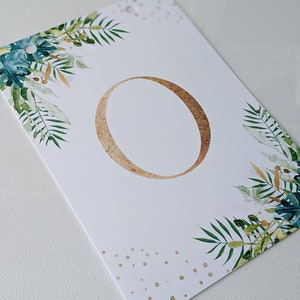 Puede incluir: Tarjeta blanca con el n&uacute;mero "0" en oro en el centro. La tarjeta est&aacute; decorada con follaje verde y dorado y lunares dorados.
