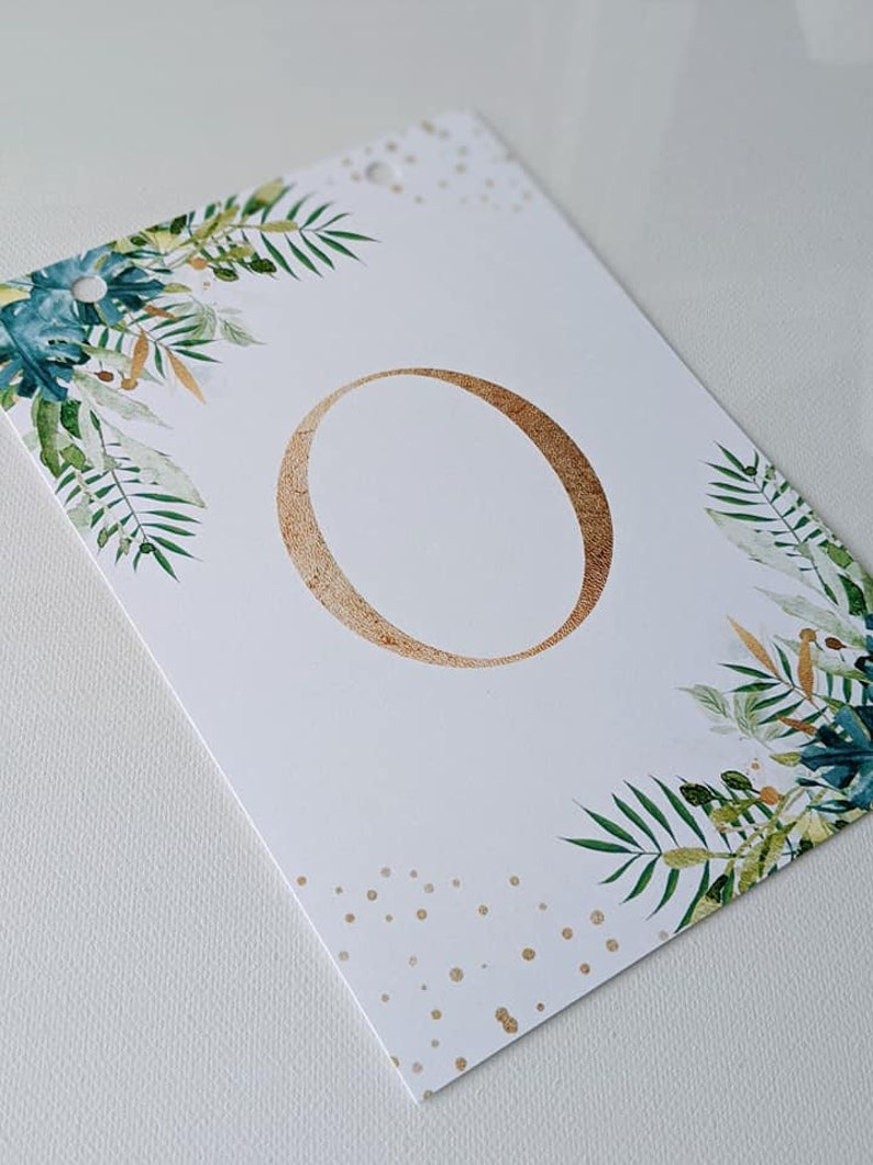 Puede incluir: Tarjeta blanca con el n&uacute;mero "0" en oro en el centro. La tarjeta est&aacute; decorada con follaje verde y dorado y lunares dorados.