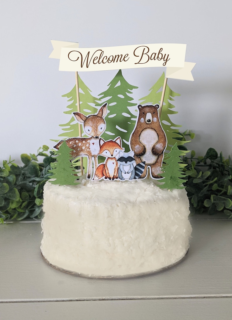 K&ouml;nnte beinhalten: Ein wei&szlig;er Kuchen mit einem Waldtier-Motiv. Der Kuchen ist mit einem Banner verziert, auf dem "Willkommen Baby" steht, sowie einem B&auml;ren, einem Reh, einem Fuchs und einem Waschb&auml;ren. Der Kuchen ist von gr&uuml;nen Bl&auml;ttern umgeben.