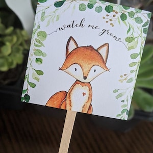 May include: A square plant marker featuring a watercolor illustration of a fox with orange fur and a heart on its chest. The text "watch me grow" is written above the fox, surrounded by a green leaf border. The marker is attached to a wooden stick.