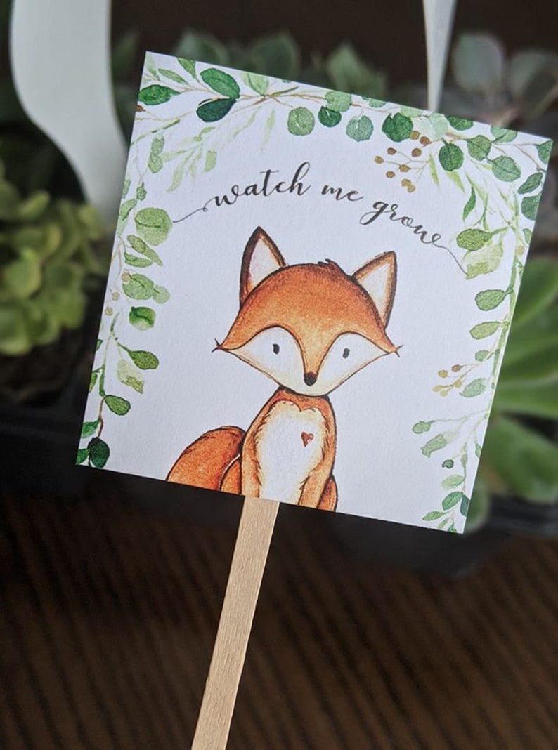 May include: A square plant marker featuring a watercolor illustration of a fox with orange fur and a heart on its chest. The text "watch me grow" is written above the fox, surrounded by a green leaf border. The marker is attached to a wooden stick.