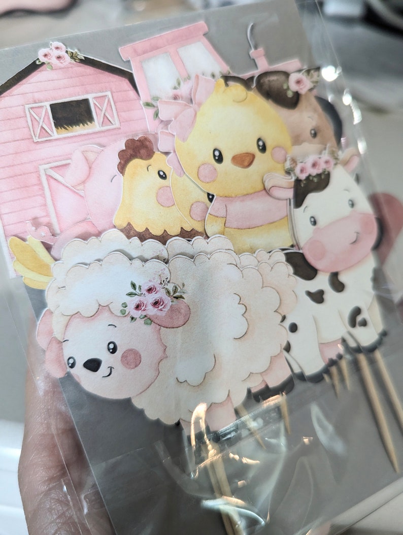 May include: A set of six pink and white farm animal cupcake toppers. The toppers feature a pig, a chick, a sheep, a cow, a barn, and a chick with a pink bow.
