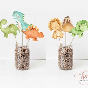 May include: A set of dinosaur themed party decorations. The decorations include a green dinosaur, a blue dinosaur, an orange dinosaur, a volcano, a lion, and a green triceratops. The decorations are arranged in two clear glass jars filled with brown paper shreds.