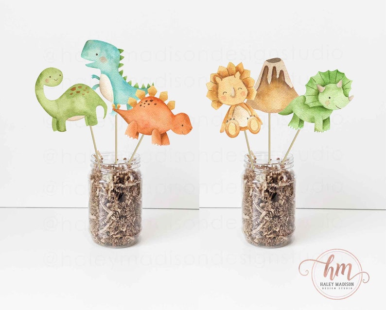 May include: A set of dinosaur themed party decorations. The decorations include a green dinosaur, a blue dinosaur, an orange dinosaur, a volcano, a lion, and a green triceratops. The decorations are arranged in two clear glass jars filled with brown paper shreds.